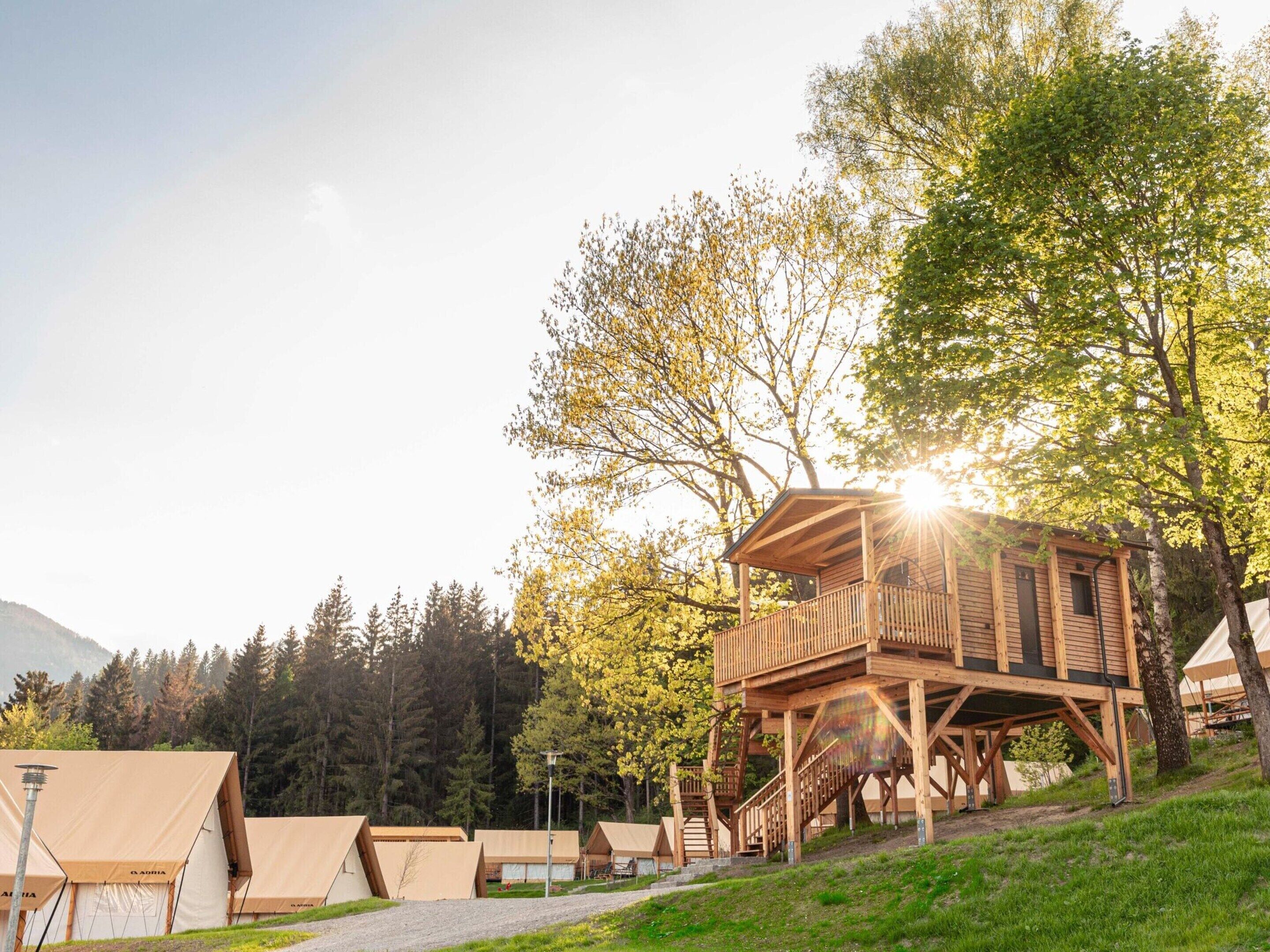 A wooden cabin with a balcony, surrounded by tents and trees, with a clear sky.