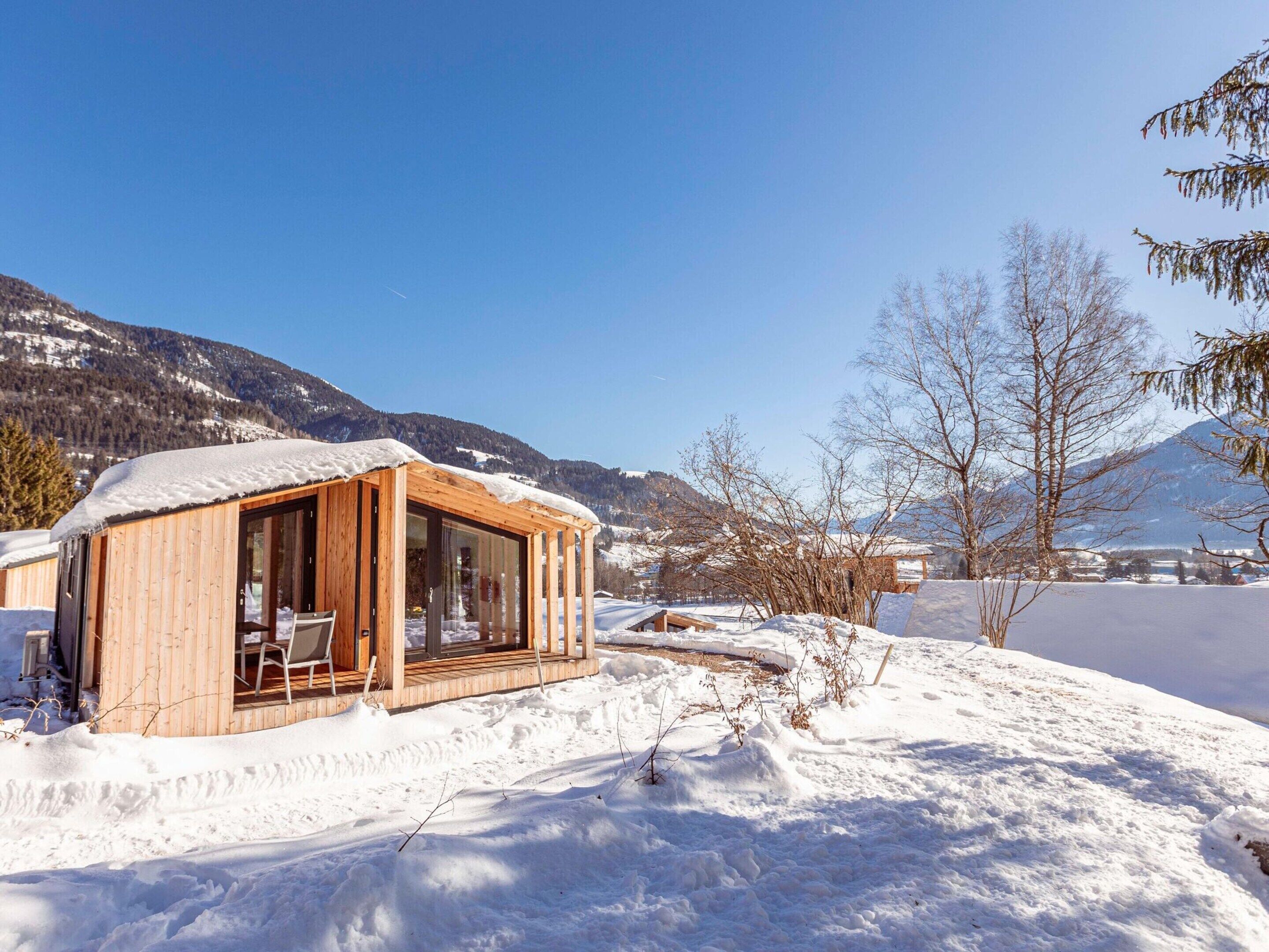 A wooden cabin with large windows, surrounded by snow-covered ground and mountains in the background.