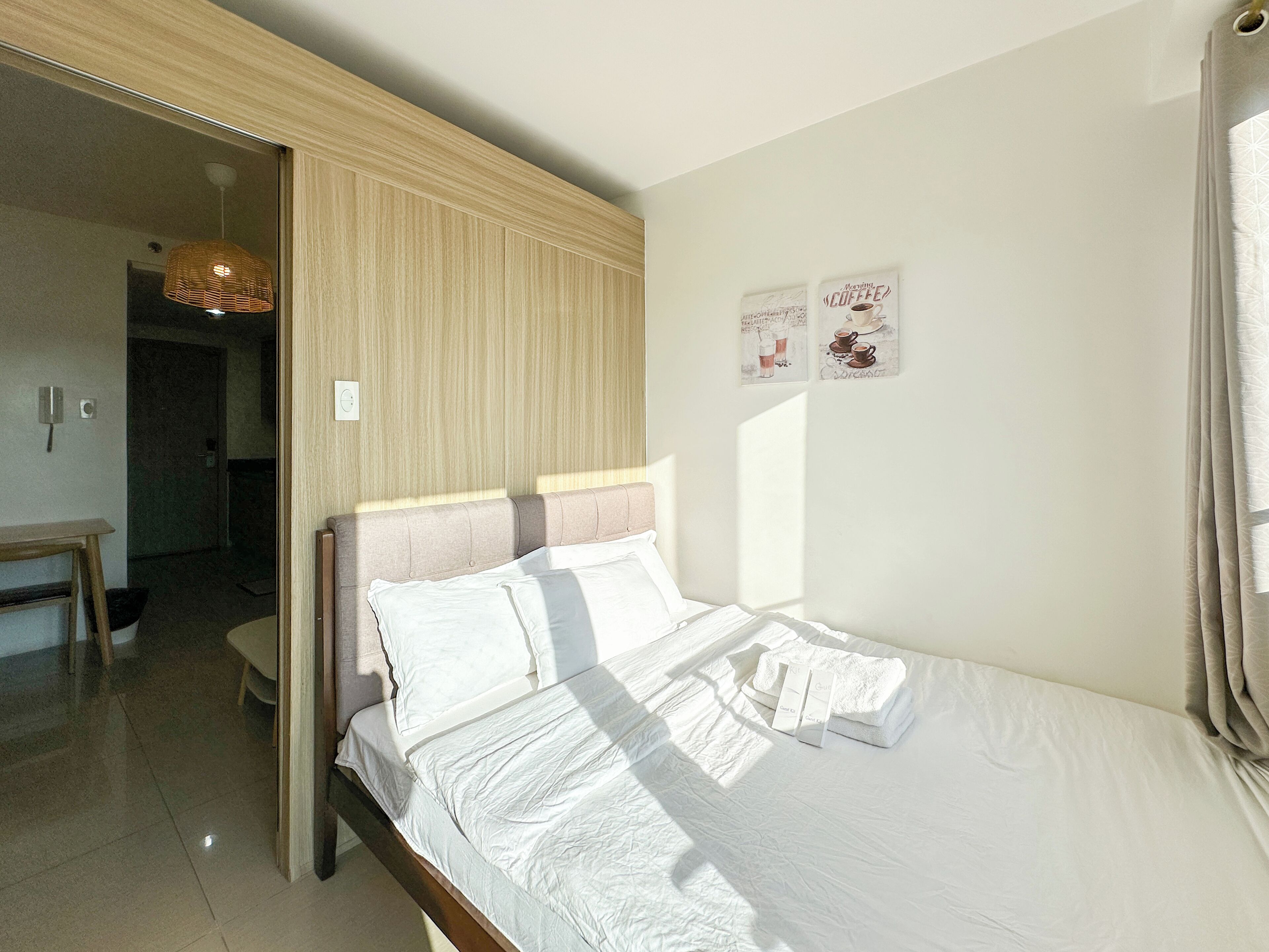 A neatly made bed with white linens, a wooden headboard, and a small bedside table. There are two framed pictures on the wall, and a door leading to another room.