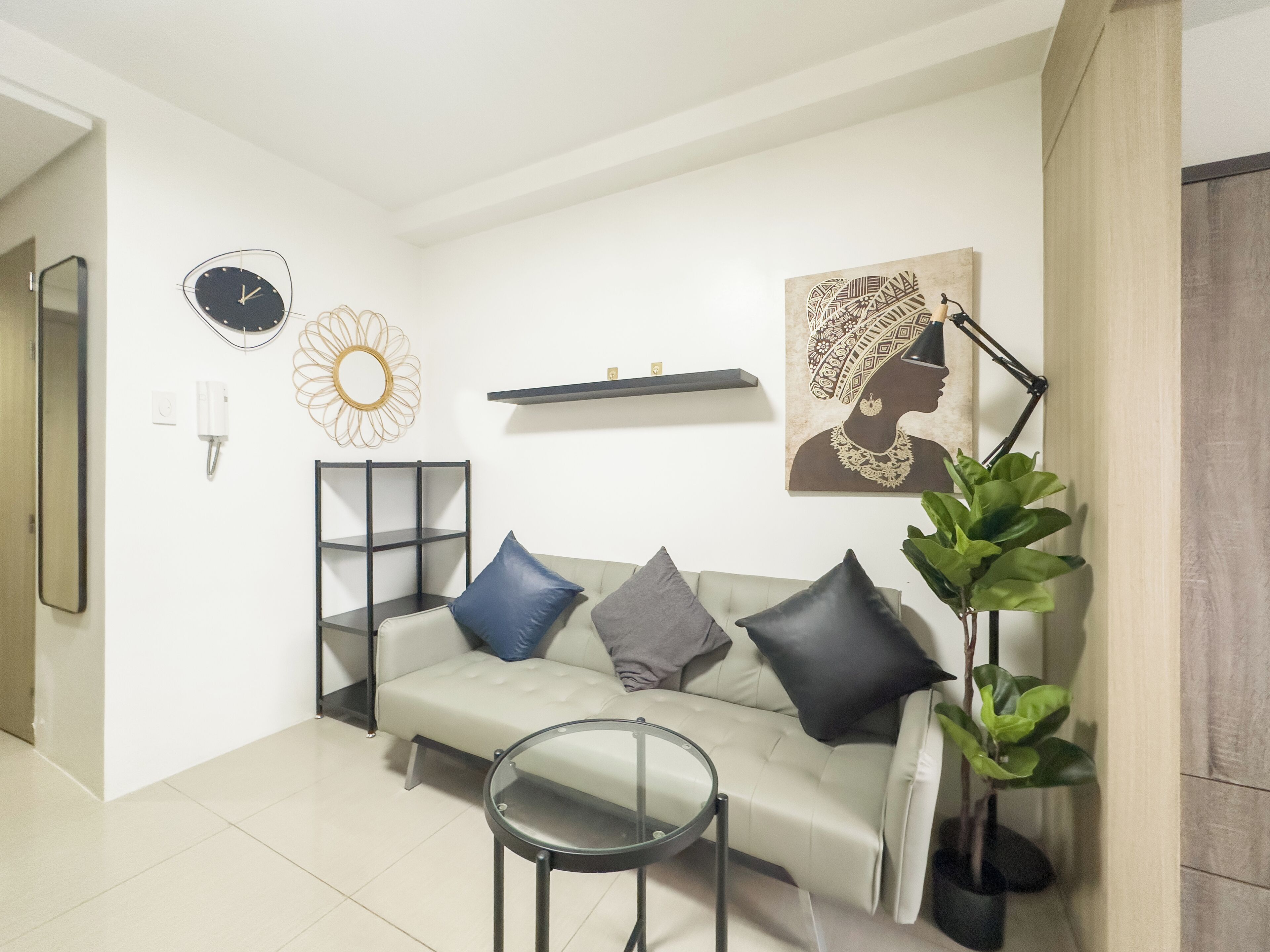 A living room with a sofa, a small round table, a floor lamp, a potted plant, a wall clock, a decorative mirror, and a shelf with a framed picture.