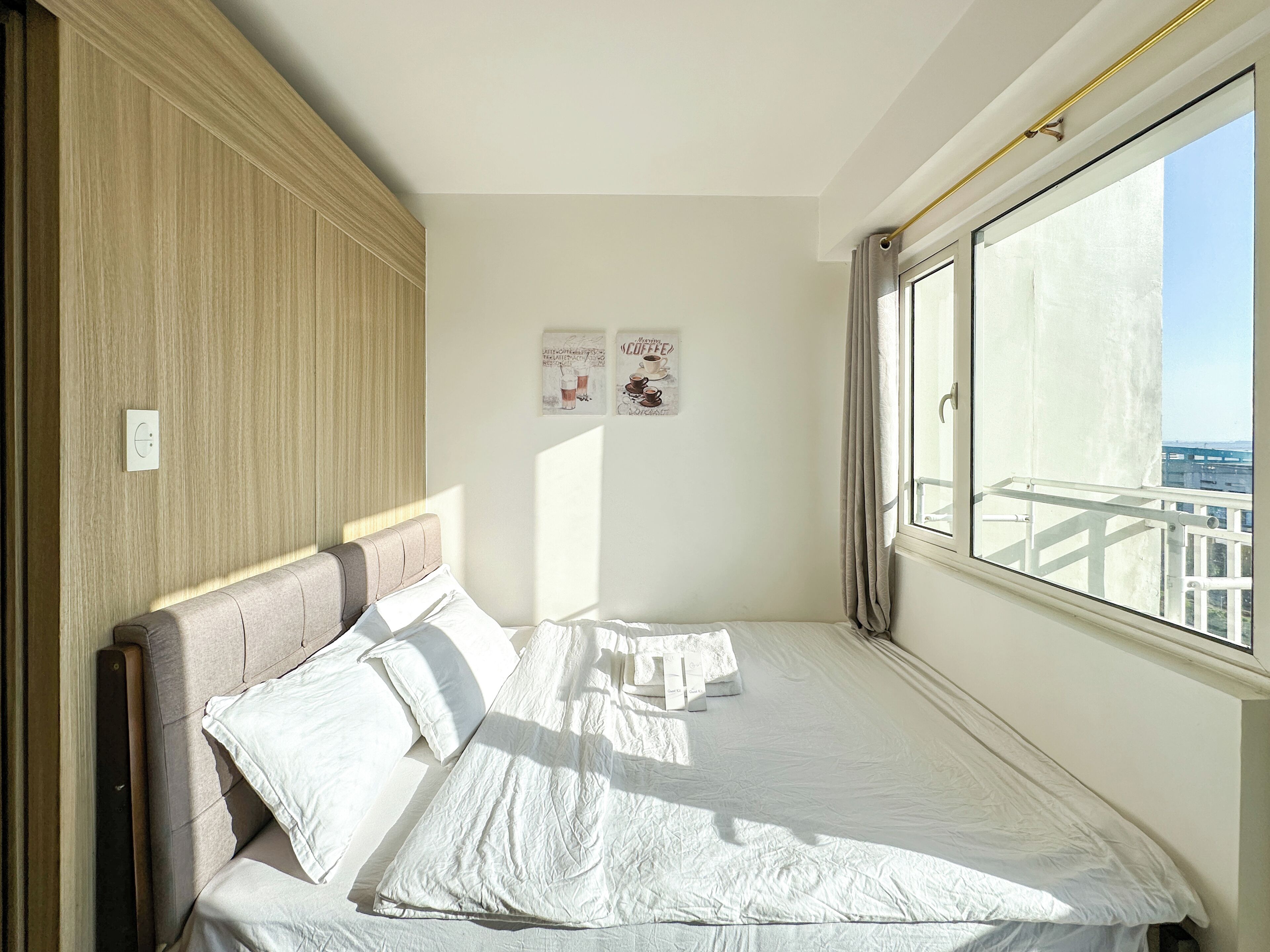 A neatly made bed with white linens, a wooden headboard, and a view of the city through a large window.