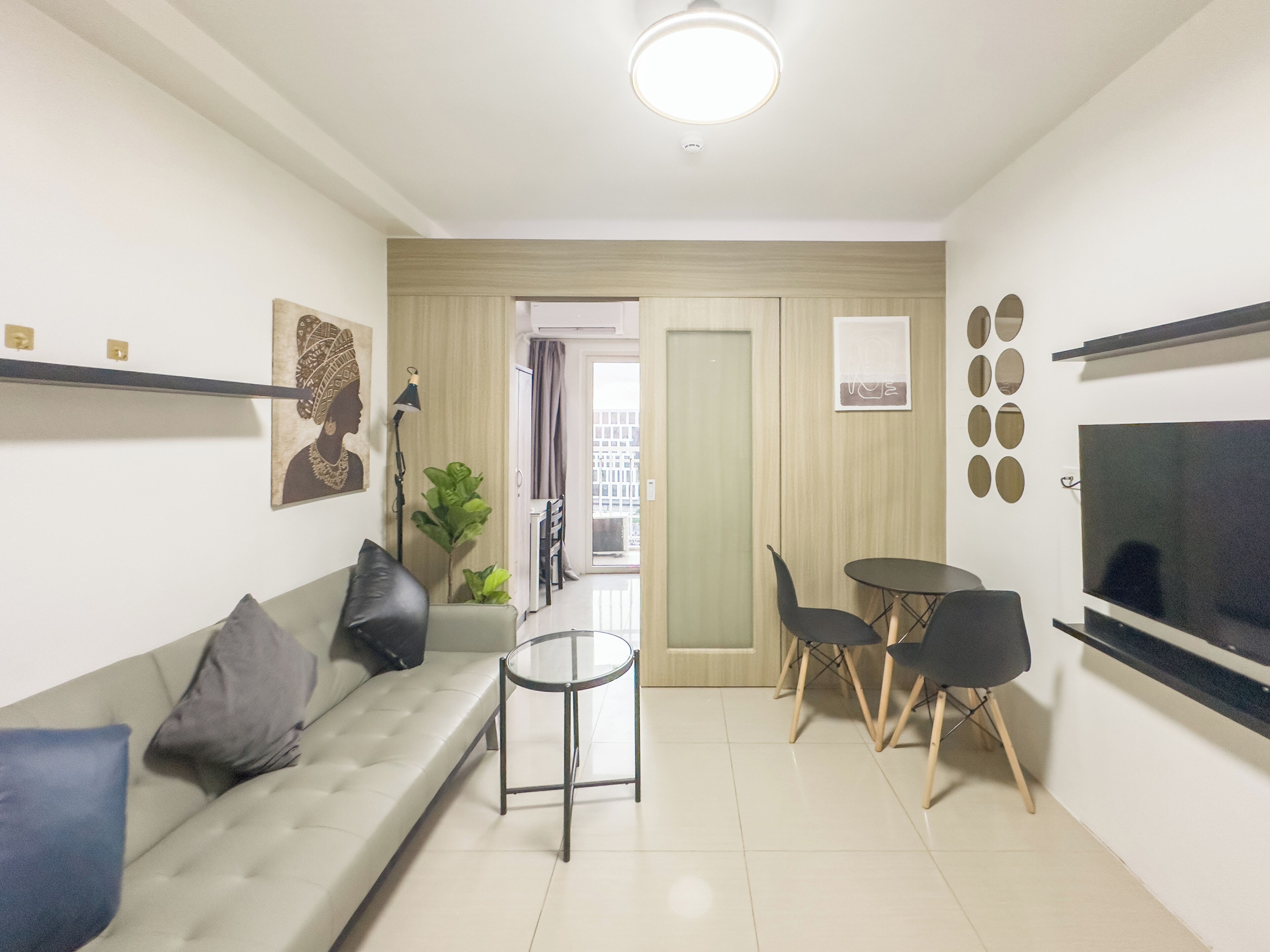 A modern living room with a sofa, a small round table, and two chairs. There is a television on the right wall and a door leading to a balcony.