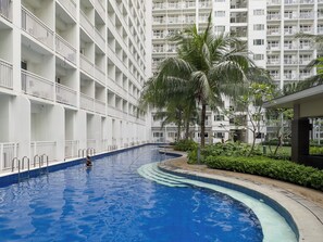 Una piscina de borde curvo, rodeada por un edificio blanco con balcones y palmeras.