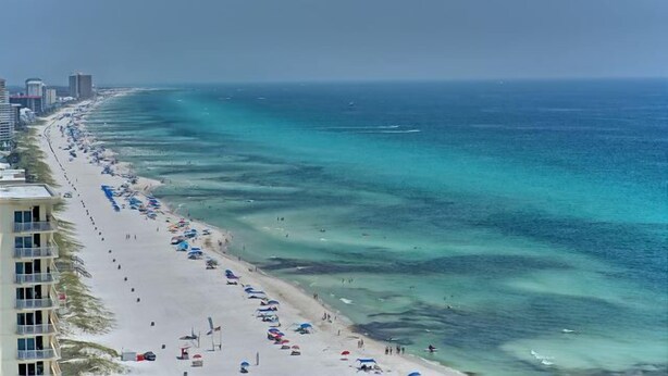 Ein Strand mit weitem Sandstreifen, klarem, blauem Wasser und einer Reihe von Gebäuden entlang der Küste.