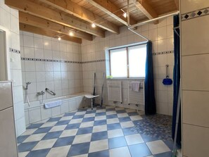 A bathroom with a checkered floor, white tiled walls, a window with a curtain, and a shower area.
