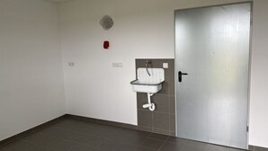 Una habitación con piso de baldosas grises, un lavamanos blanco instalado en la pared, una puerta y un detector de incendios rojo.