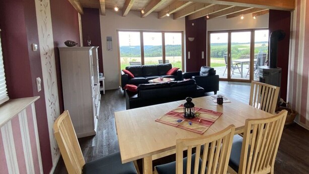 Amplia sala de estar con techo de madera, una mesa de comedor con sillas, un sofá y vistas al exterior a través de amplios ventanales.