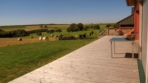 Una terraza de madera con vistas a un paisaje agrícola, que incluye pacas de heno y árboles.