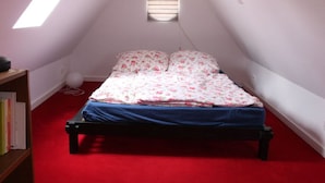 A small, cozy attic bedroom with a sloped ceiling, a single bed with floral bedding, a red carpet, and a small window with blinds. - Cozy Apartment on the Canal (Friedrichstadt)