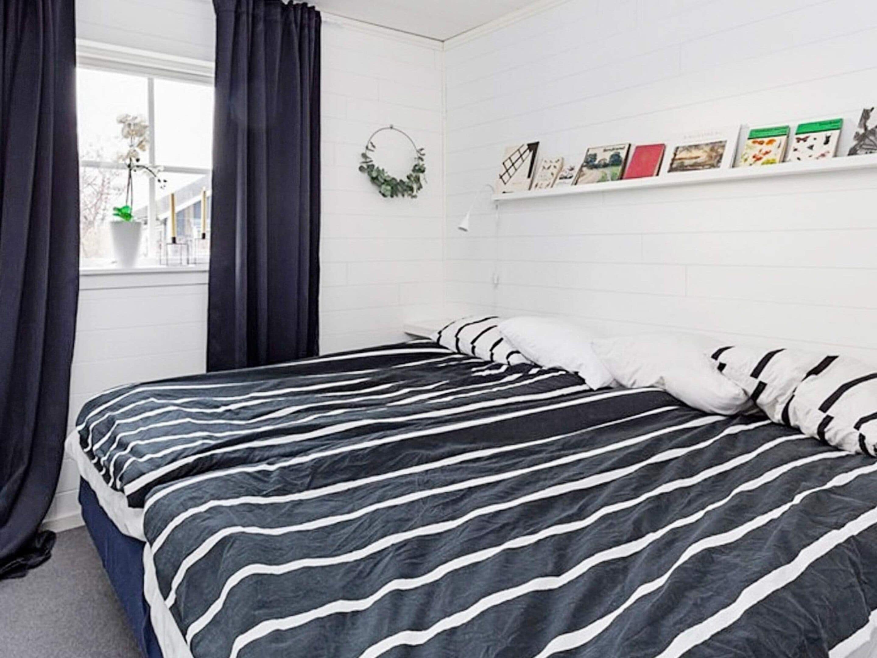 Un dormitorio con cama, una ventana con cortinas, una repisa en la pared con libros y una guirnalda.