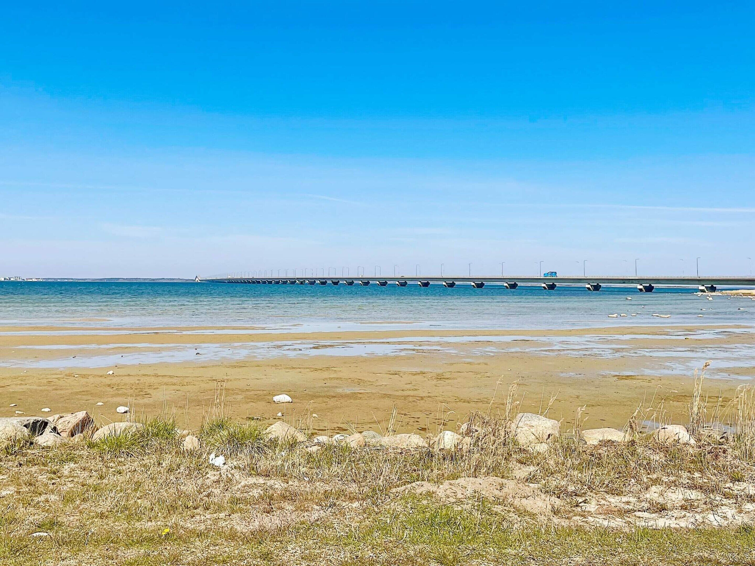Ein Strand mit einer langen Mole, die ins Wasser führt, einem klaren Himmel und einem sandigen Ufer mit vereinzelter Vegetation.