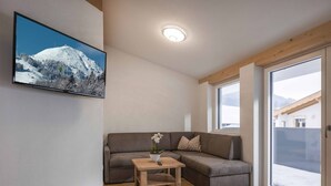 Un salon moderne avec un plancher en bois, un canapé d’angle gris, une petite table en bois ornée d’un vase de fleurs et une grande fenêtre donnant sur une montagne enneigée.
