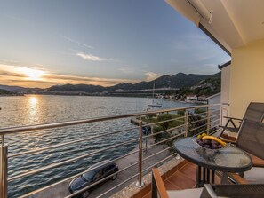 A balcony with a view of a lake and mountains, a table with fruits, and a car parked nearby. - Apartments Ema-comfort Two Bedroom Apartment With Sea View (Ploce)