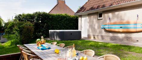 An outdoor dining area with a long wooden table, wicker chairs, and a buffet tray with food and drinks.
