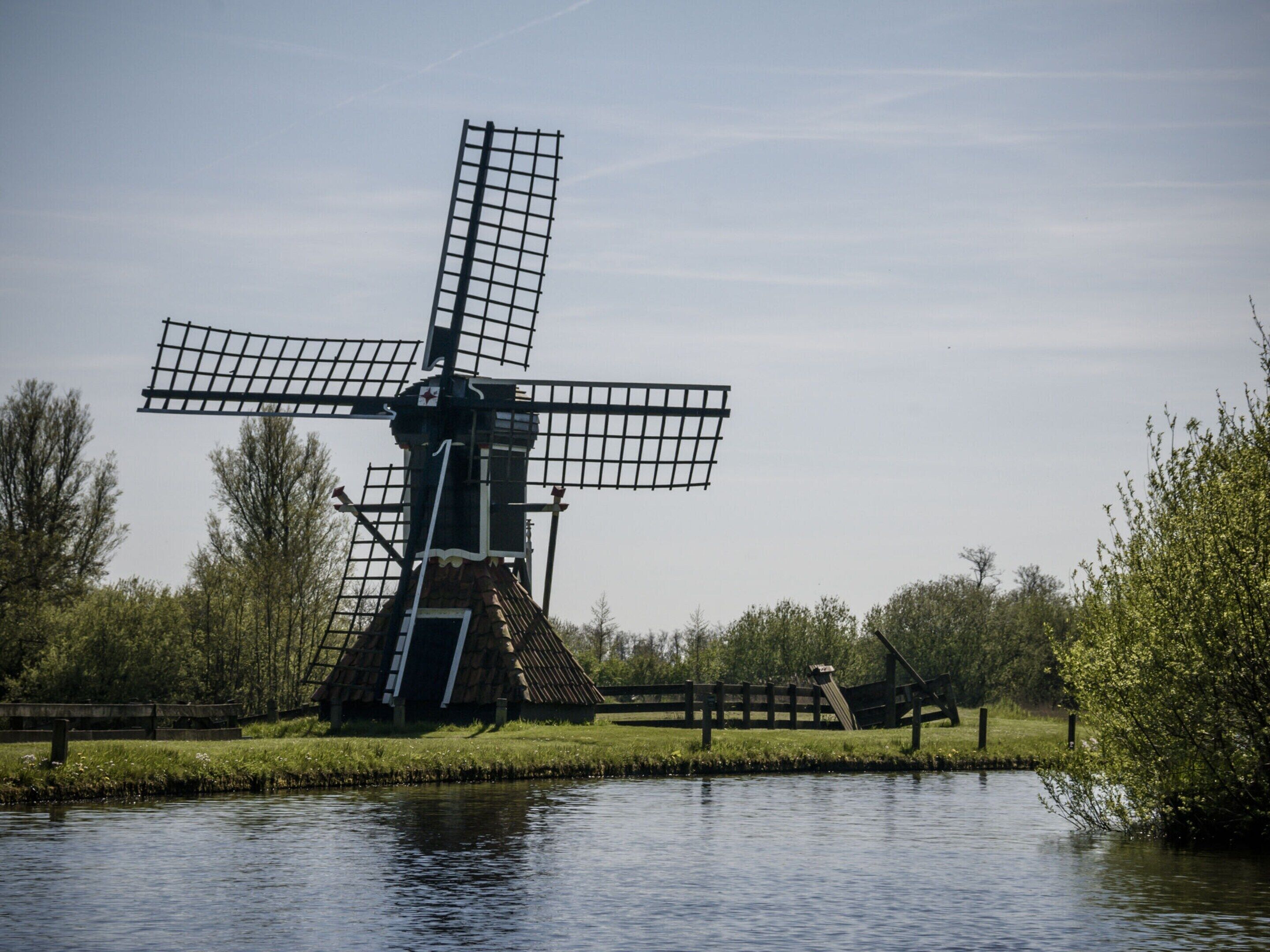 Un moulin à vent traditionnel, situé au bord d’une rivière paisible, entouré de verdure et délimité par une clôture en bois.