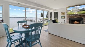 A spacious living room with a large window overlooking a body of water, a fireplace, and a dining area with blue chairs.