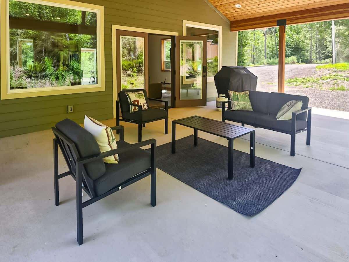 An outdoor patio with a black metal table and two black sofas with cushions, a wooden ceiling, and a view of greenery through large windows.