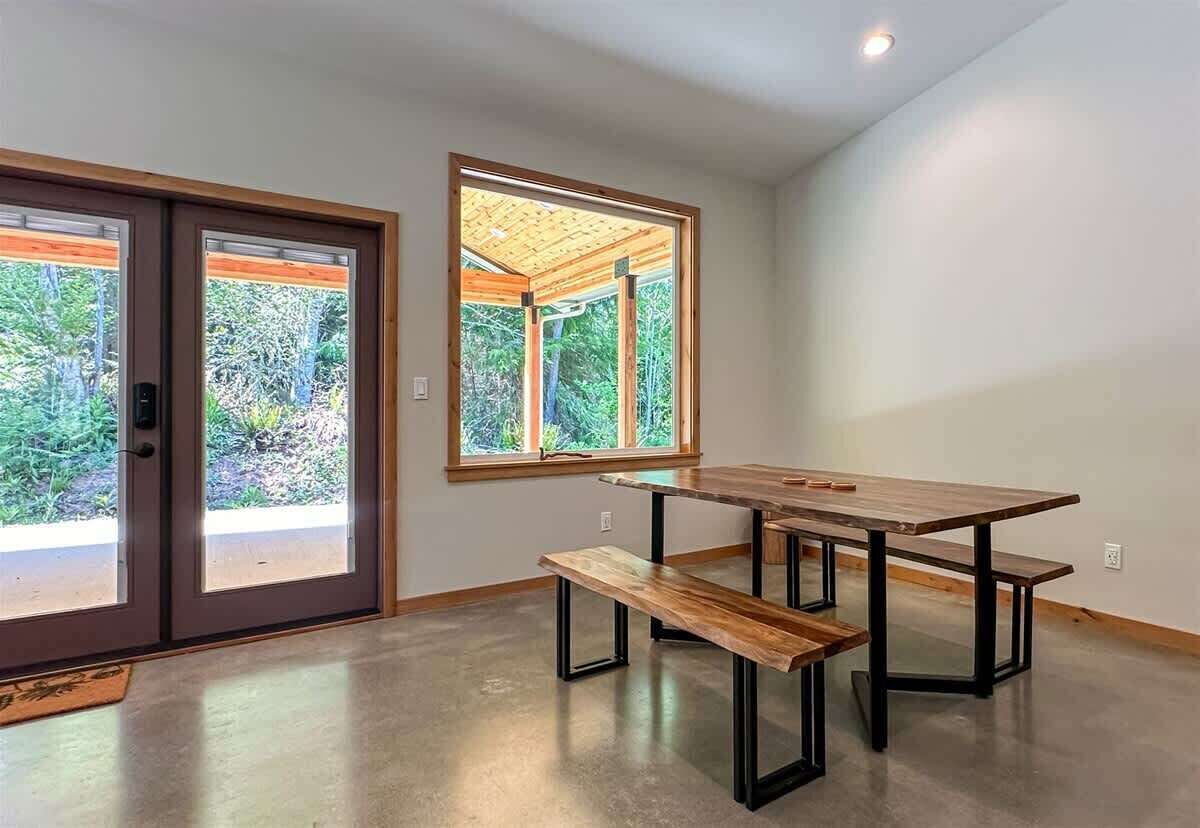 A modern interior with a wooden table and benches, large windows with a view of greenery, and a ceiling with recessed lighting.