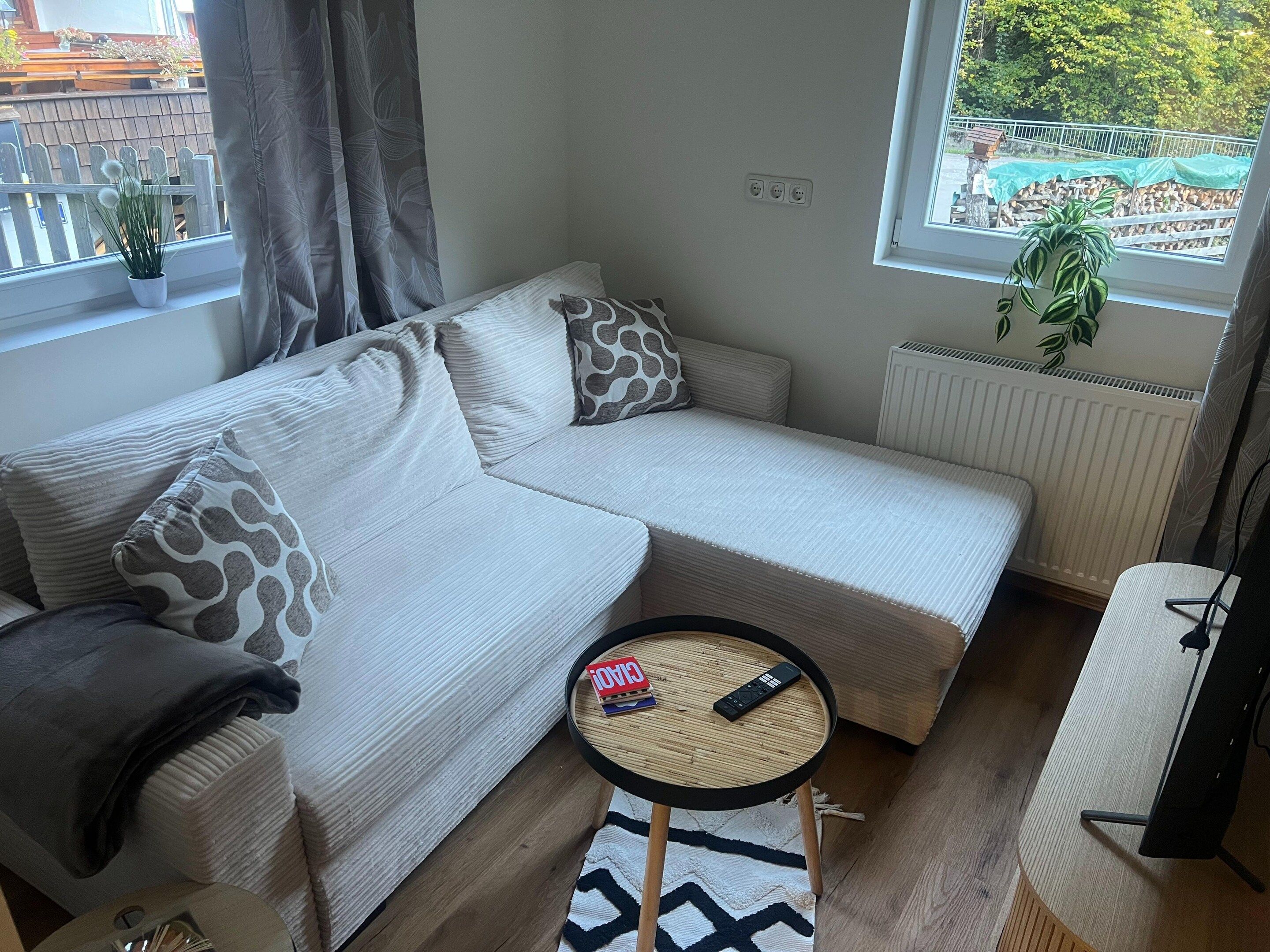 A modern living room with a sectional sofa, a small round table, and a view of a garden through a window.