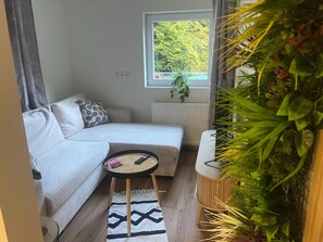 A cozy room with a sofa, a small round table, a window with a view of greenery, and a radiator. - Apartment in Dorfzentrum Near Lake (Feld am See)