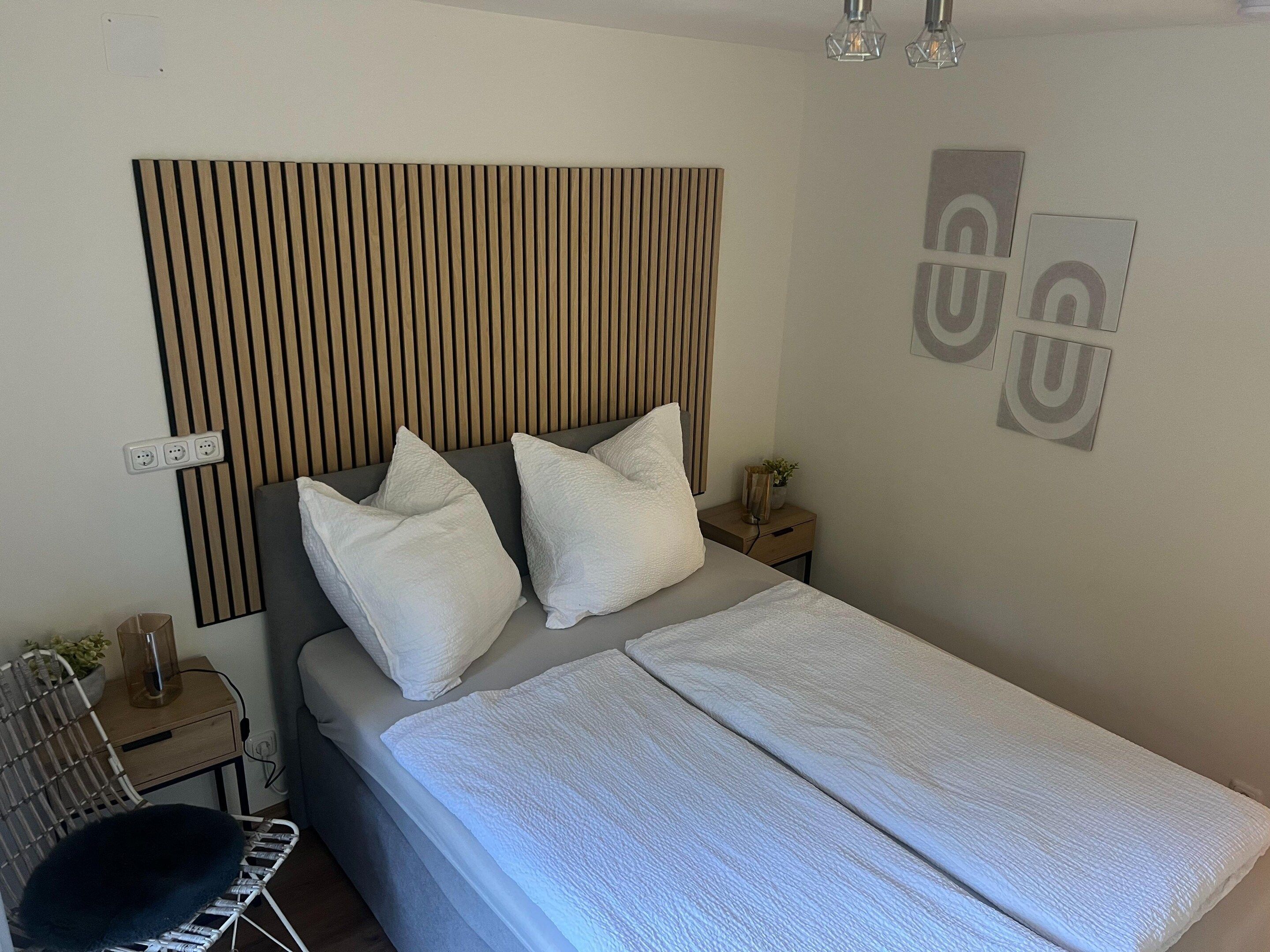A bedroom with a bed, pillows, a bedside table with a lamp, a wall-mounted power outlet, and a wall decoration.
