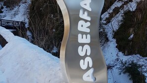 Une enseigne métallique sur laquelle est inscrit le mot « JASERFALL » dans une police de caractères stylisée, le tout se détachant sur un paysage enneigé avec des arbres et un pont en bois en arrière-plan.