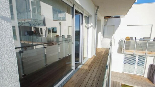Un balcon moderne avec des garde-corps en verre, une terrasse en bois et un mur extérieur blanc.