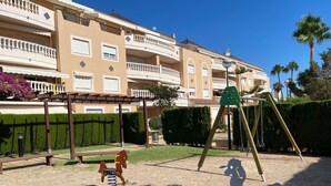 Ein Spielplatz mit Schaukeln und einer Wippe vor einem mehrstöckigen Gebäude mit Balkonen und einem gepflegten Garten.