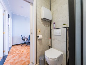 Et badeværelse med toilet, vægmonteret klimaanlæg, en lille hylde med toiletartikler og flisegulv.
