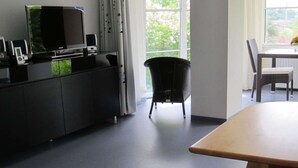 Une pièce moderne avec un plancher gris foncé, une table en bois, une chaise noire, une chaise en osier, un téléviseur et un meuble.