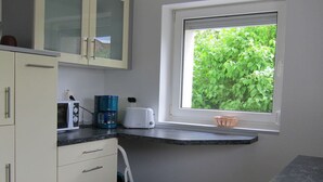 Une cuisine compacte équipée d’un micro-ondes, d’un grille-pain et d’une machine à café sur le plan de travail, d’une petite fenêtre donnant sur un espace verdoyant et d’un meuble blanc avec des tiroirs.
