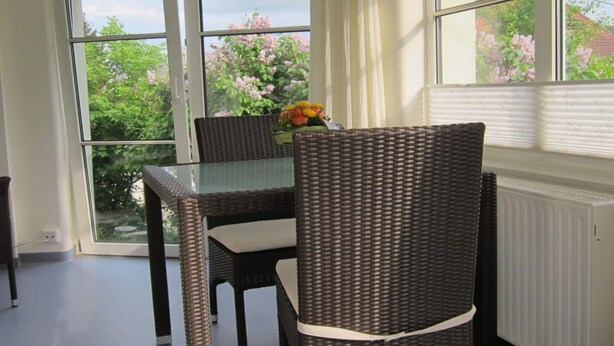 Un coin repas avec des meubles en osier, une table en verre et une vue sur la verdure grâce à de grandes fenêtres.
