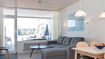 Una sala de estar moderna con un sofá gris, una mesa de centro redonda y una mesa de comedor con sillas. Hay lámparas colgantes, un ventanal y vistas al exterior.