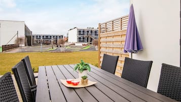 A wooden table with chairs, a potted plant, and a purple umbrella in a courtyard with buildings in the background.