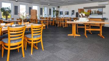 A spacious dining area with wooden tables and chairs, large windows, and indoor plants.