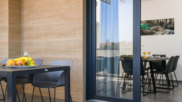 A modern dining area with a wooden wall, a glass door leading to an outdoor seating area, and a table set with fruits and glasses. - Villa Dora - Apt 3 "grijs" (Pula)