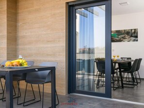 A modern dining area with a wooden wall, a glass door leading to an outdoor seating area, and a table set with fruits and glasses. - Villa Dora - Apt 3 "grijs" (Pula)