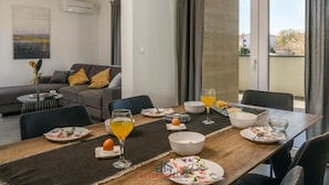 A dining area with a table set for breakfast, a sofa in the background, and a view through the window. - Villa Dora - Apt 3 "grijs" (Pula)