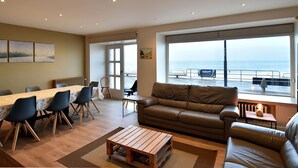A spacious living room with a large window offering a view of the sea, a dining area with a table and chairs, and a comfortable seating area with a sofa and armchairs. - Giannina 0001 Located at the Beach (De Haan)