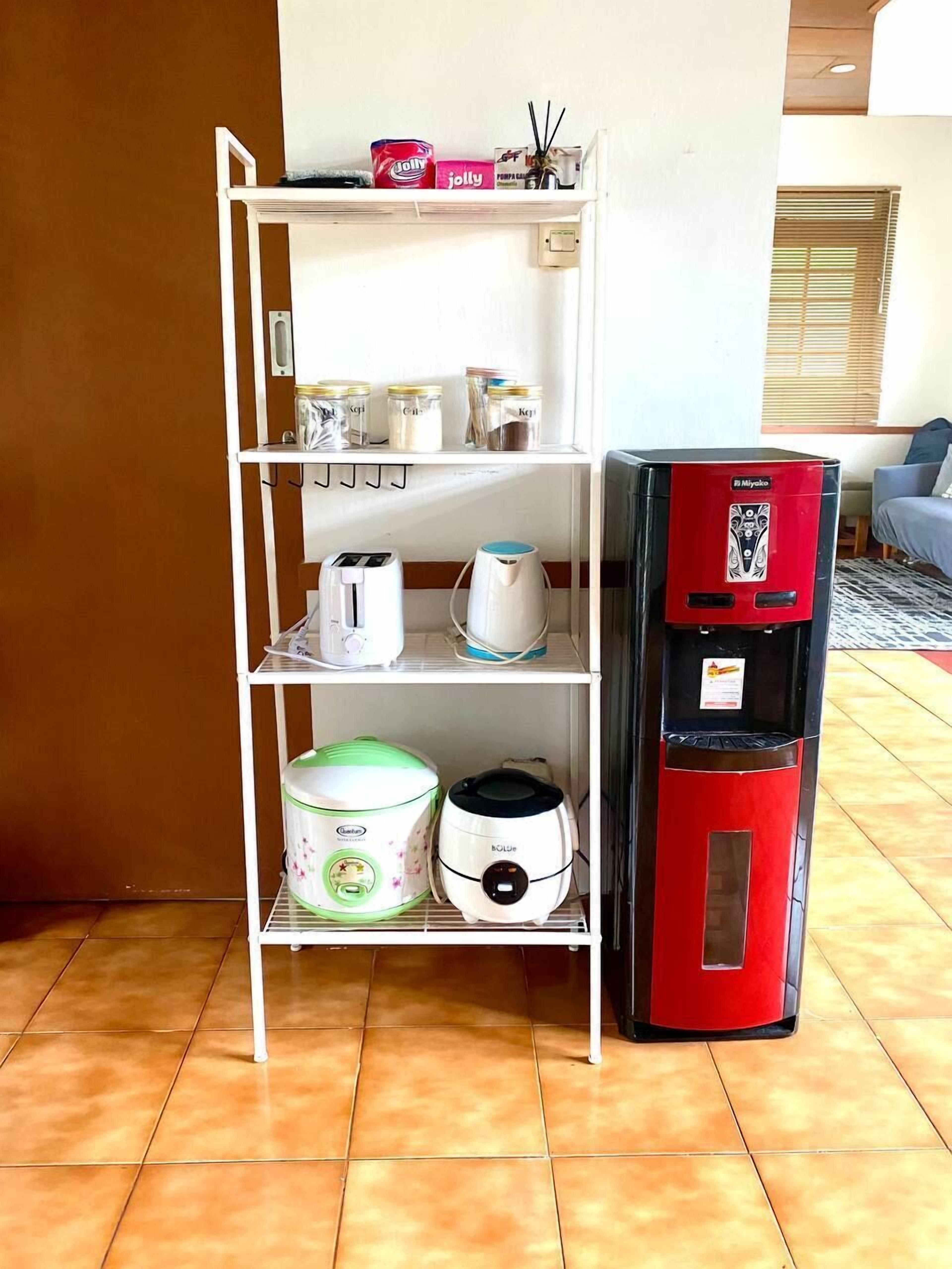 Un área de cocina con un estante metálico blanco que contiene electrodomésticos y frascos de almacenamiento. Un dispensador de agua rojo se encuentra a la derecha.