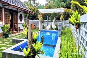 Una piscina con borde de baldosas, rodeada de plantas en macetas y una pérgola.