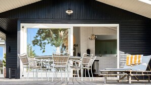 Een overdekt terras met een tafel, stoelen en een lounge stoel, met zicht op het interieur door een glazen deur.
