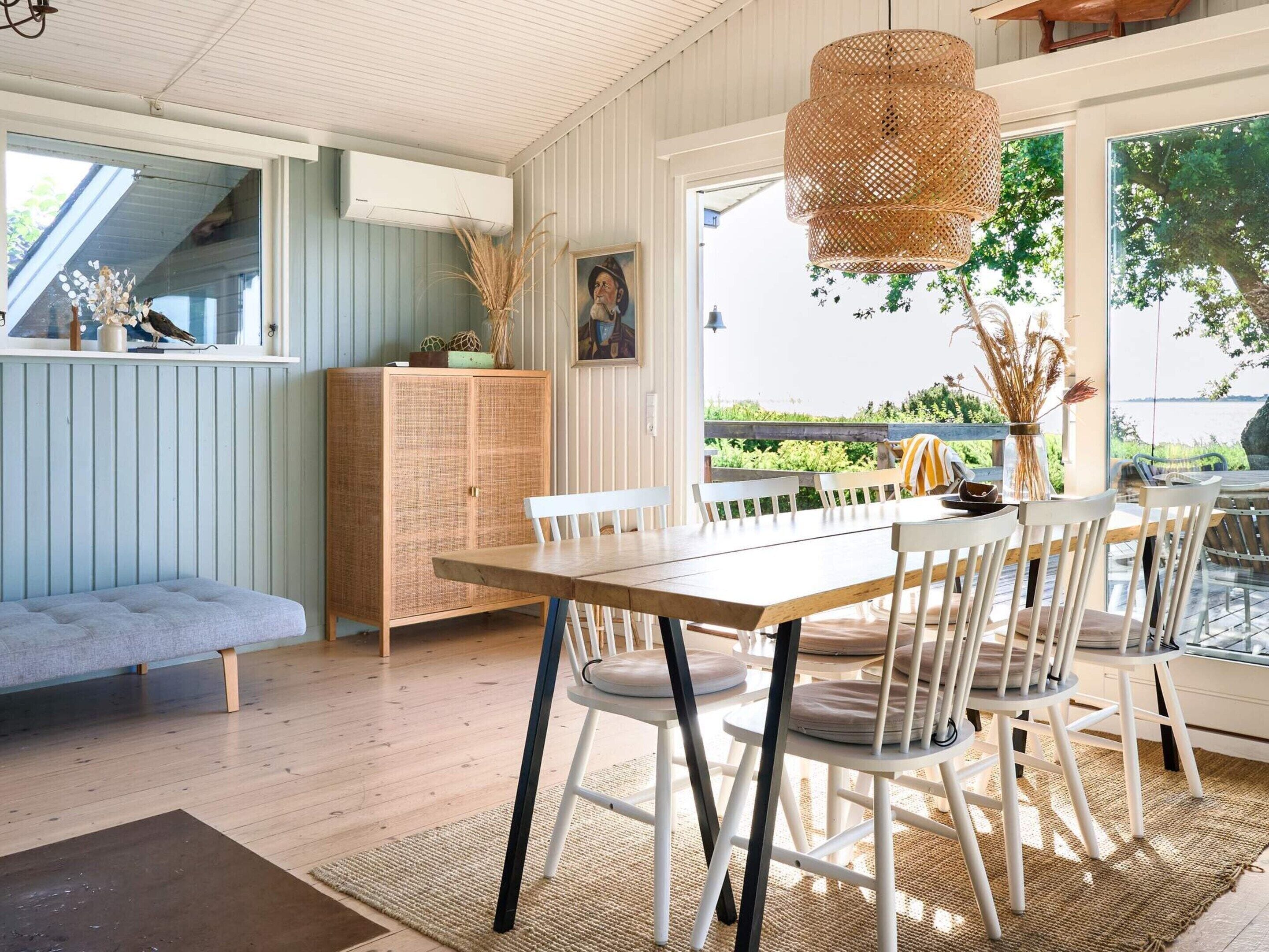 A dining area with a wooden table and white chairs, a woven pendant light, and a view of greenery outside.