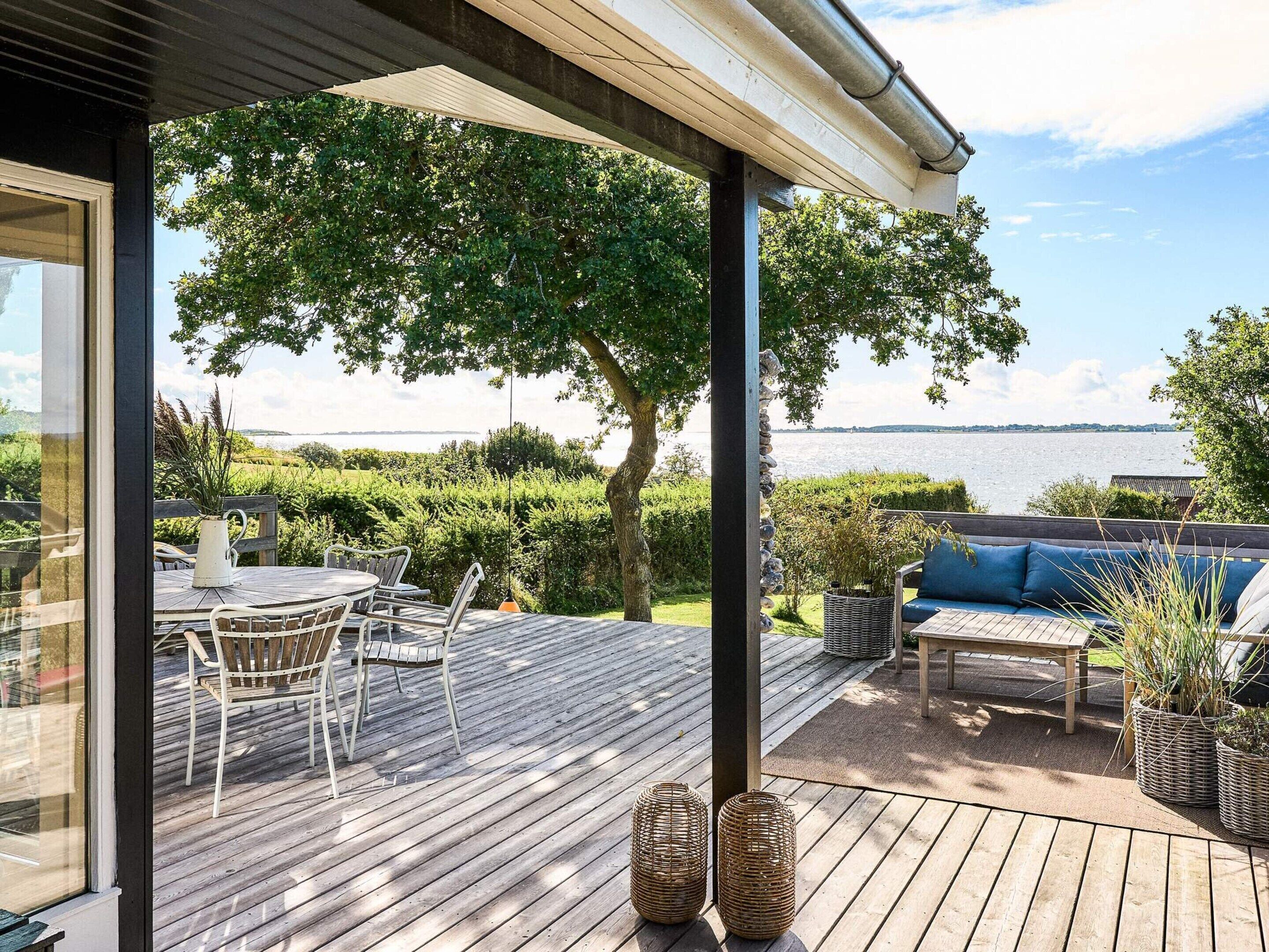 Una terraza de madera con mobiliario exterior, vista al agua y un árbol.