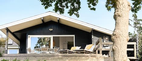 A modern house with a large deck, outdoor seating, and a tree in the foreground.
