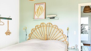 Quarto com cabeceira de vime, cama com colcha branca, luminária fixada na parede e uma obra de arte emoldurada sobre a cama.