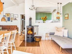 A cozy living room with a wood-burning stove, a sofa, a dining table with chairs, and a kitchen area with green cabinets and a window. - 6 Person Holiday Home in Faaborg (Faaborg)