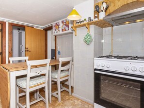 A compact kitchen with a gas stove, white chairs, a wooden table, and a yellow hanging lamp. - 2 Person Holiday Home in Snedsted (Snedsted)
