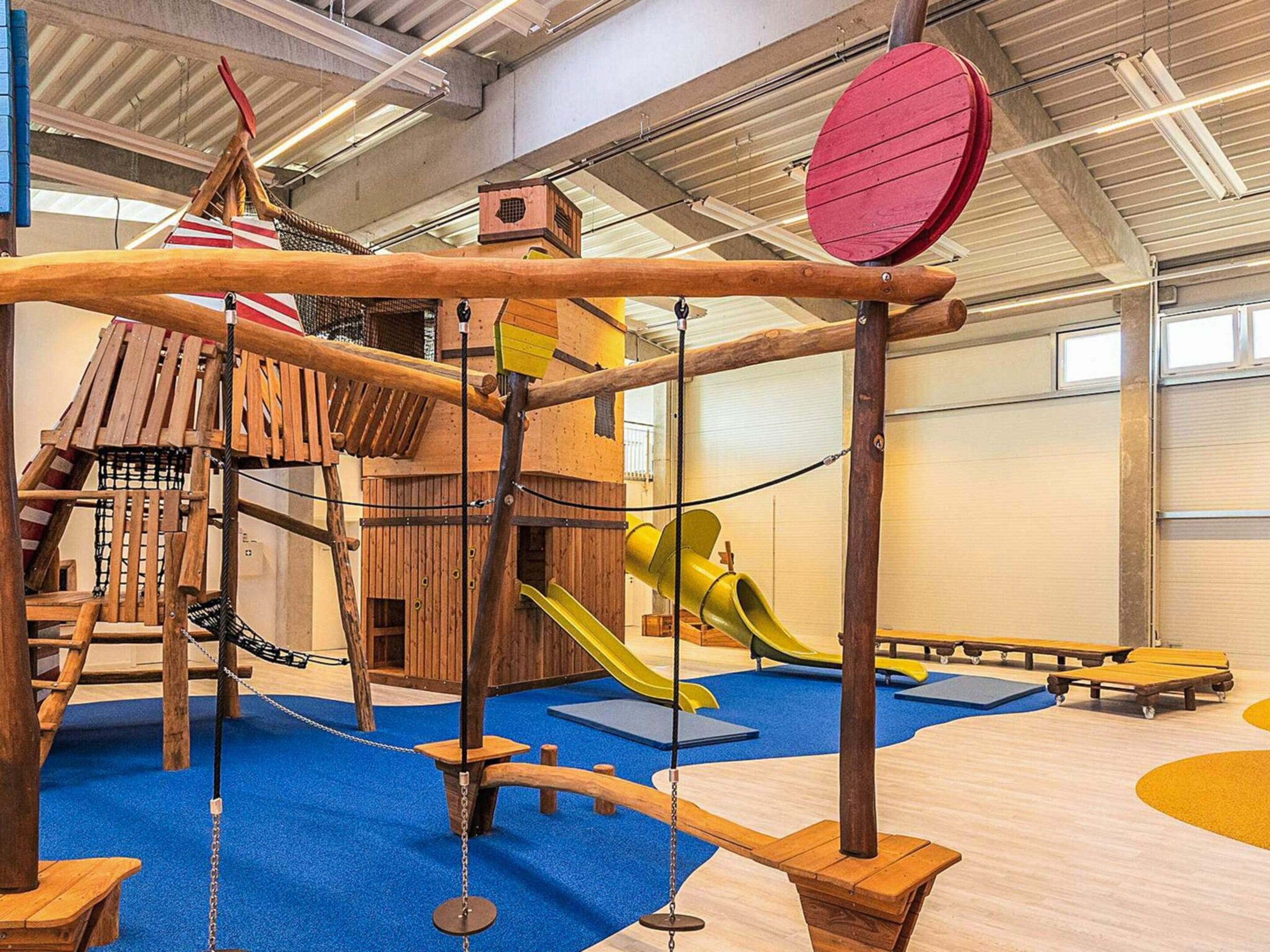 A children's play area with a wooden climbing structure, slides, and soft play mats.