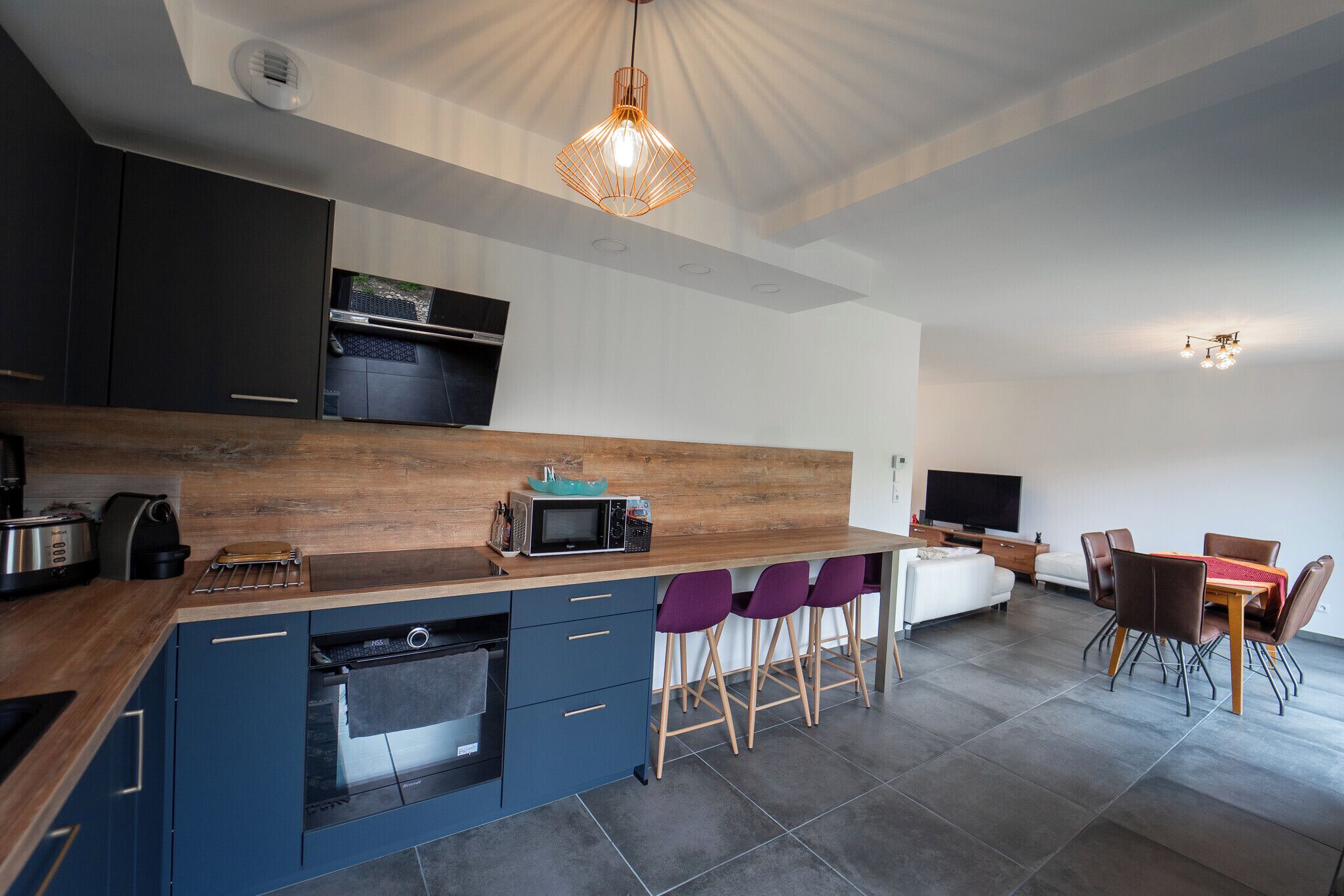 Una cocina moderna con armarios oscuros, encimera de madera y horno empotrado. Hay sillas moradas y una mesa de comedor en la zona de comedor contigua.